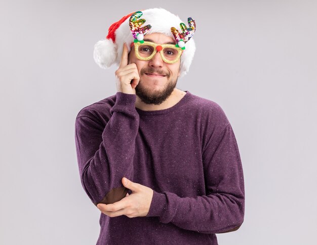 보라색 스웨터와 흰색 배경 위에 유쾌 하 게 서 웃 고 카메라를보고 재미있는 안경을 쓰고 산타 모자에 행복 한 젊은 남자