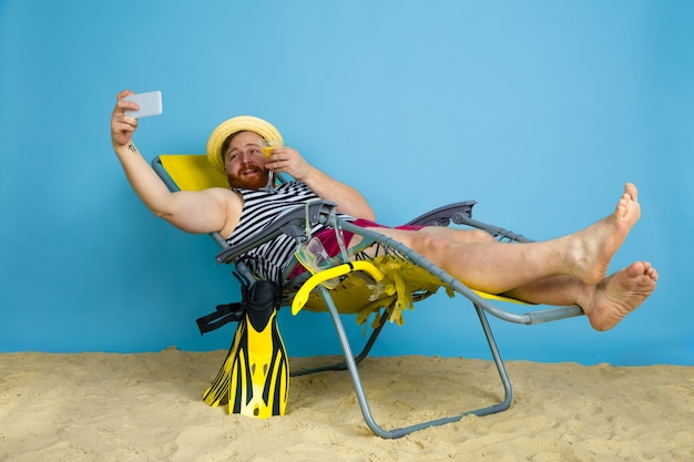 Felice giovane uomo che riposa, prende selfie, bevendo cocktail su sfondo blu studio. concetto di emozioni umane, espressione facciale, vacanze estive o fine settimana. freddo, estate, mare, oceano, alcol.