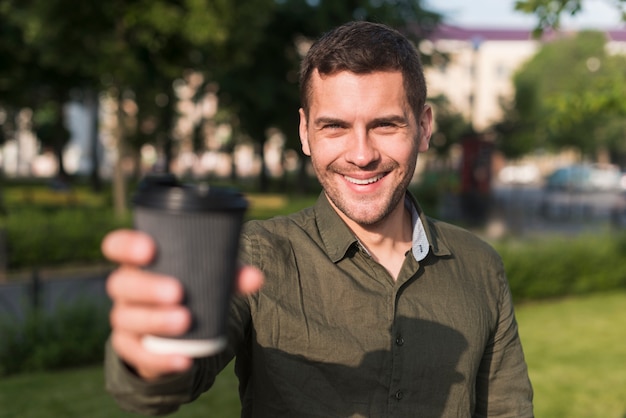 公園で使い捨てのコーヒーカップを示す幸せな若い男