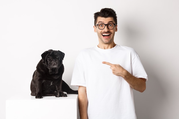 Happy young man showing his cute dog, pointing finger at black pug and smiling, standing over white background