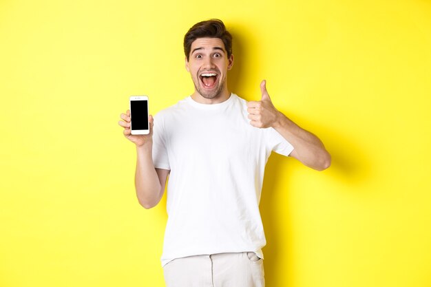 Happy young man showing thumb up and mobile phone screen, recommending application or internet website, standing over yellow background