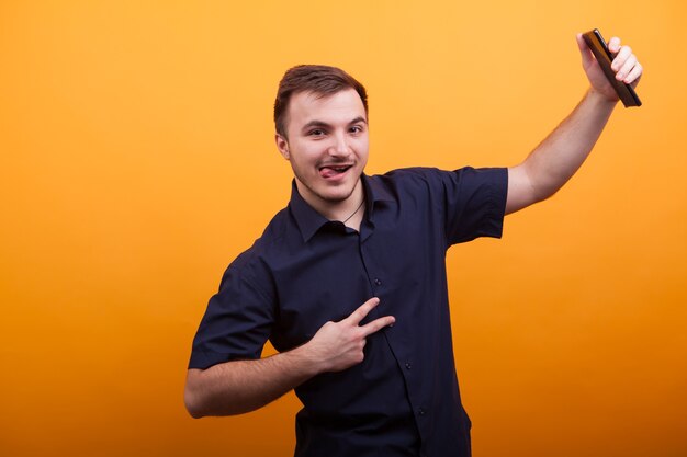 Happy young man showing victory sign and holding mobile phone over yellow backdround. Funny young man