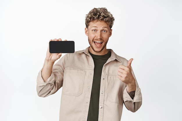 Happy young man shows horizontal phone screen and thumbs up recommending smartphone app pleased with awesome application white background