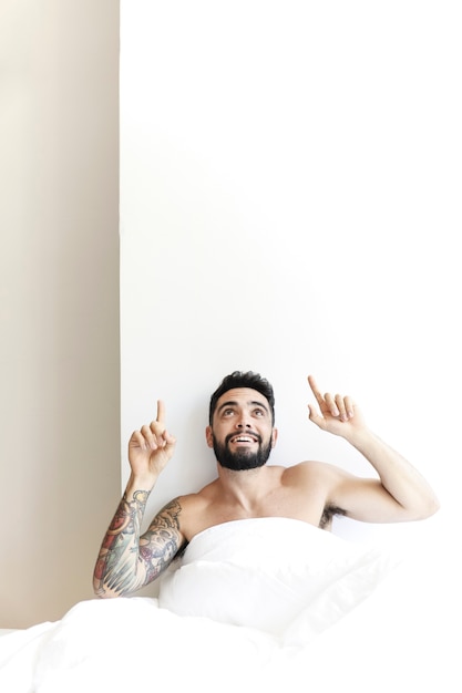 Happy young man sitting on bed pointing upwards