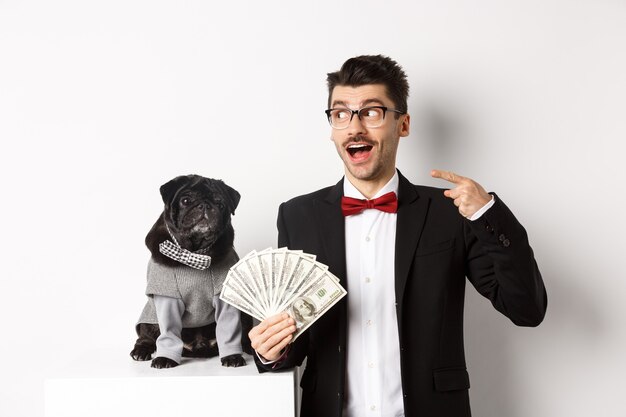 Happy young man in suit earn money with his dog. Guy rejoicing, holding dollars and pointing left, black pug in costume staring at camera, white background.