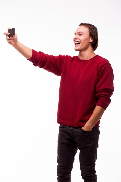 Счастливый молодой человек, делающий автопортретную фотографию через смартфон, изолированный на белой стене