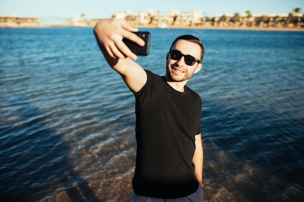 Счастливый молодой человек в отпуске смеется на пляже, принимая селфи в солнцезащитных очках на море