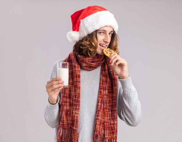 Happy  young man wearing christmas santa hat with warm scarf around his neck holding glass of milk eating cookie standing over white  wall