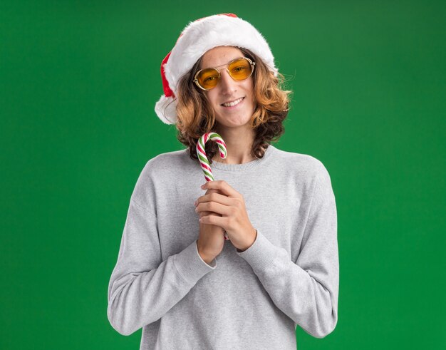 Счастливый молодой человек в рождественской шляпе санта-клауса и желтых очках, держащий конфету, смотрит в камеру с улыбкой на лице, стоя на зеленом фоне