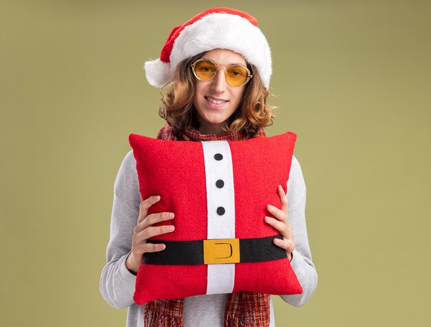 Happy young man wearing christmas santa hat and yellow glasses with warm scarf around his neck holding christmas pillow  with smile on face  standing over green wall