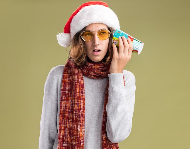 緑の背景の上に立って混乱しているように見える彼の耳の上にカラフルな紙コップを保持している彼の首の周りに暖かいスカーフとクリスマスサンタ帽子と黄色いメガネを身に着けている幸せな若い男