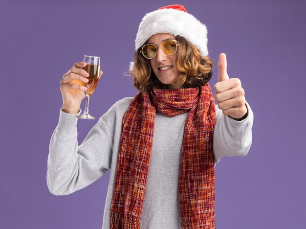 クリスマスのサンタの帽子と黄色いメガネを身に着けている幸せな若い男は、首に暖かいスカーフを持って、紫色の壁の上に立って親指を見せて笑っているシャンパンのガラスを持っています