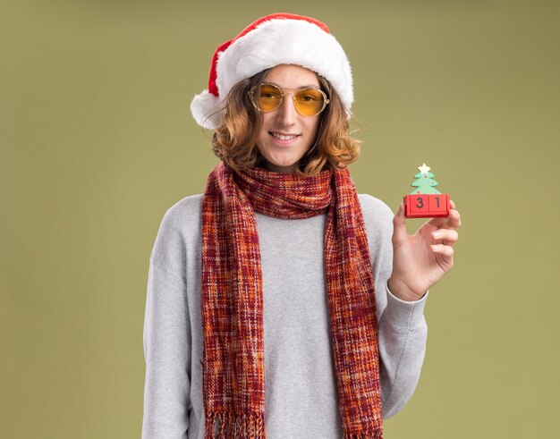 녹색 벽 위에 서있는 얼굴에 미소와 함께 새해 날짜와 장난감 큐브를 들고 그의 목에 따뜻한 스카프와 함께 크리스마스 산타 모자와 노란색 안경을 쓰고 행복 한 젊은 남자