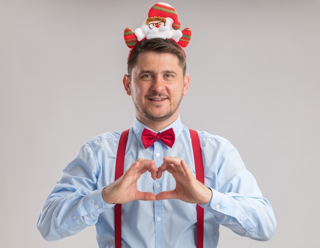 Felice giovane uomo che indossa bretelle farfallino in cerchio con santa guardando la telecamera facendo gesto di cuore con le dita sorridendo allegramente in piedi su sfondo bianco