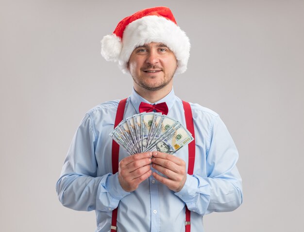 Happy young man wearing suspenders bow tie in santa hat holding cash looking at camera with smile on face standing over white background