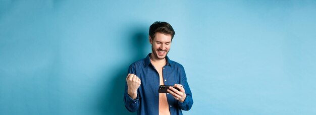 Happy young man winning in video game on mobile phone looking satisfied at smartphone screen and say