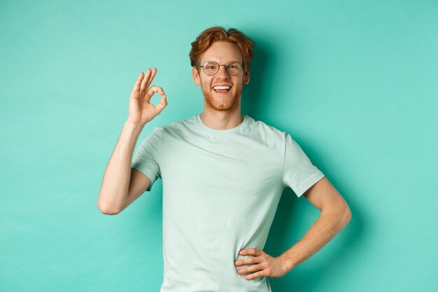 Happy young man with red hair and beard, wearing glasses and t-shirt, smiling satisfied and showing ok sign, say yes, approve and agree, standing over turquoise background