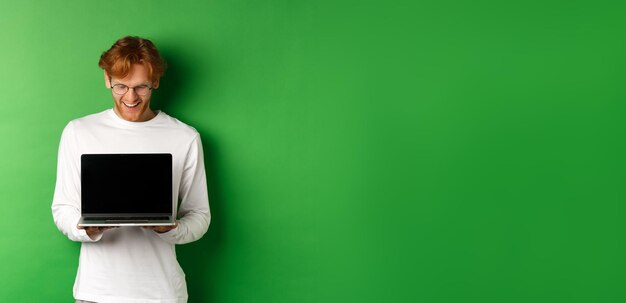 Free photo happy young man with red hair showing blank laptop screen and looking pleased smiling while staring