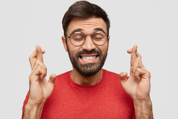 Happy young man with stubble, crosses fingers, keeps eyes closed, dressed in red t shirt