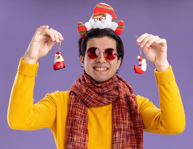 Happy young man with warm scarf around neck in yellow turtleneck and glasses with funny rim on head holding christmas toys  with smile on face standing over purple wall