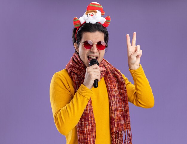 Happy young man with warm scarf around neck in yellow turtleneck and glasses with funny rim on head holding microphone singing showing v-sign standing over purple wall