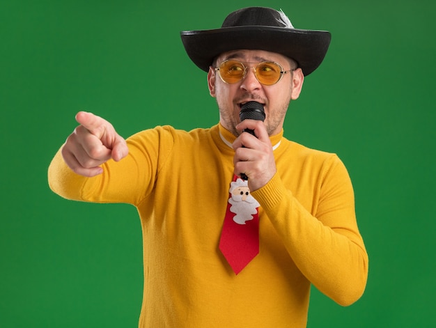 Free photo happy young man in yellow turtleneck and glasses with funny red tie holding microphone pointing with index figner to the side standing over green wall