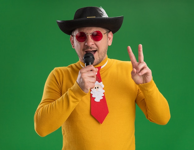 Free photo happy young man in yellow turtleneck and glasses with funny red tie holding microphone singing showing v -sign standing over green wall