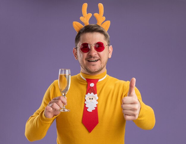 Счастливый молодой человек в желтой водолазке и красных очках, в забавном красном галстуке и оправе с оленьими рогами, держит бокал шампанского, показывая вверх большие пальцы руки, стоя на фиолетовом фоне