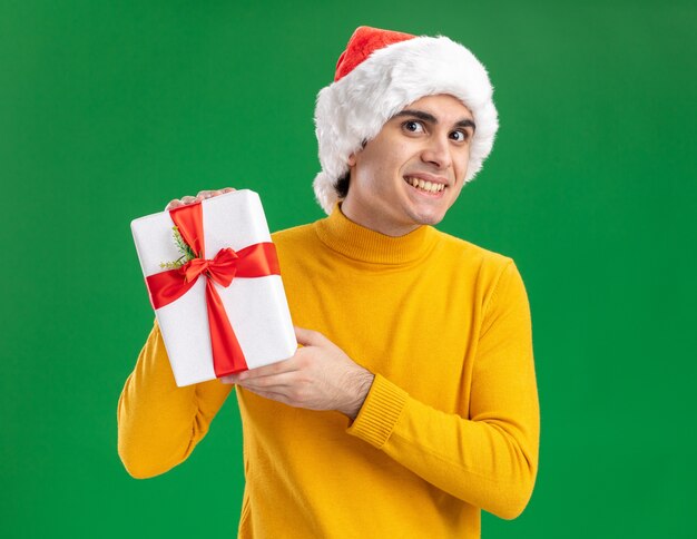 緑の背景の上に元気に立って笑顔のカメラを見てプレゼントを保持している黄色のタートルネックとサンタ帽子の幸せな若い男