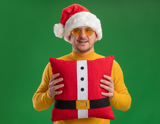 Happy young man in yellow turtleneck and santa hat wearing glasses holding red funny pillow looking at camera with smile on face standing over green background