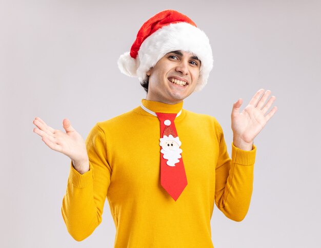 노란색 터틀넥과 흰색 배경 위에 유쾌하게 서있는 카메라를보고 재미있는 넥타이와 산타 모자에 행복 한 젊은 남자