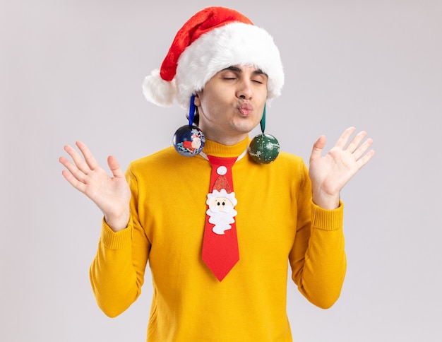 黄色いタートルネックとサンタの幸せな若い男が彼の耳にクリスマスボールを持って陽気で面白い目を閉じて白い背景の上に立っている