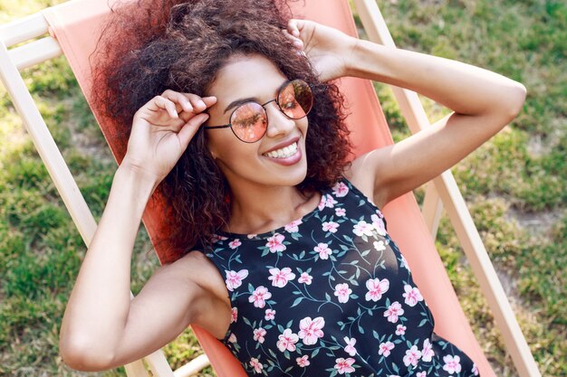 Счастливая молодая смешанная восторженная женщина с удивительными вьющимися волосами отдыхает на шезлонге на зеленой лужайке в парке
