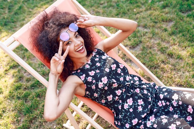 Happy young mixed rave woman with amazing curly hairs relaxing on chaise-longue  on green lawn in park