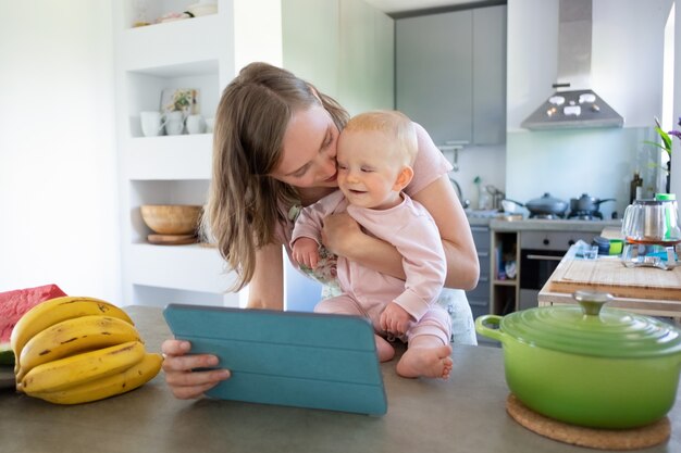 幸せな若いママと赤ちゃんの娘が台所で一緒に料理をしながらビデオ通話にタブレットを使用しています。育児や家庭料理のコンセプト