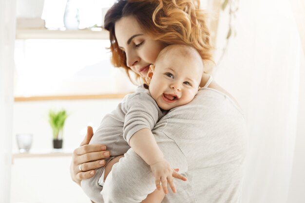 Счастливая молодая мама держит драгоценного маленького ребенка и нежно обнимает его маленькое тело. Малыш радостно смеется и смотрит в камеру большими серыми глазами.