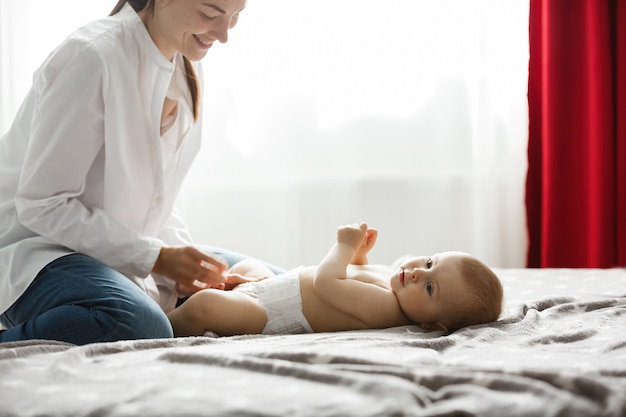 Happy young mom in light bedroom smiles and dressing her precious baby boy for a morning walk in park. Family concept..