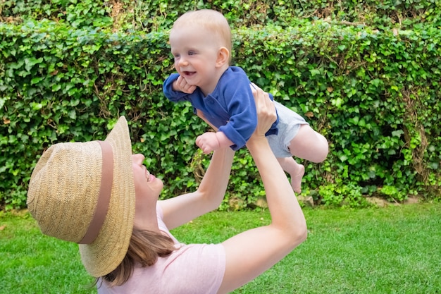 Счастливая молодая мать в шляпе, играя с младенцем в саду и смеясь. Милый ребенок в синей рубашке, глядя с открытым ртом