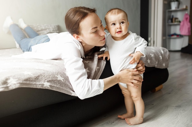 Happy young mother lying on bed in sleep room trying to kiss her precious baby. Little boy standing in front of mom , smiling and looking aside.