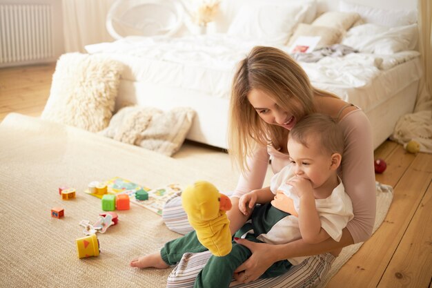 幸せな若い母親が遊んでいて、かわいい小さな子供が床で遊んでいます。黄色いアヒルのおもちゃを保持している寝室のカーペットの上に座っている愛らしい幼児に座っている金髪の女性。母性と育児の概念