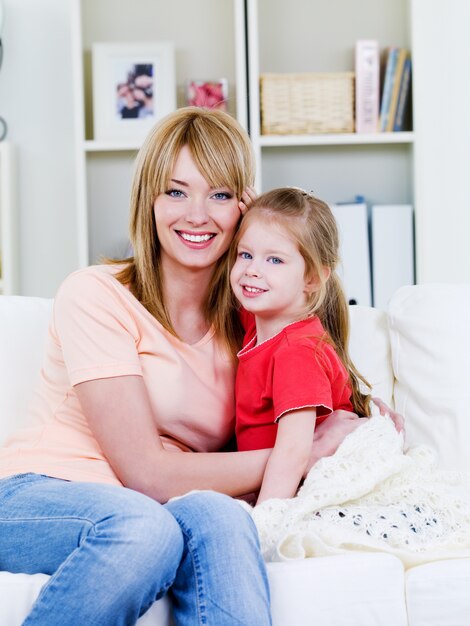 Happy young  mother  sitting on the sofa and embracing her loving little daughter - indoors