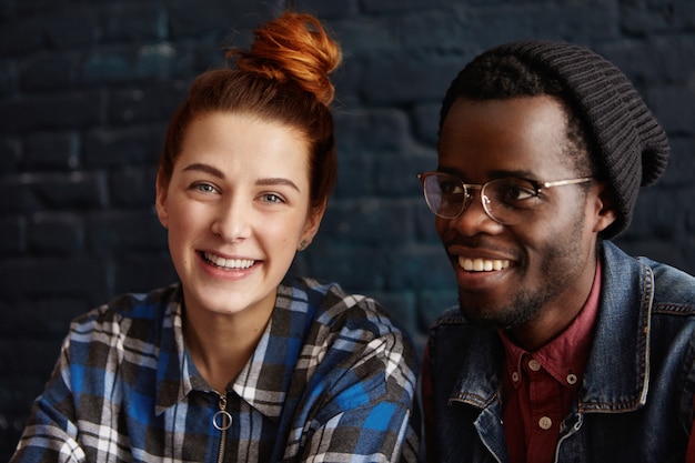 Free photo happy young multiethnic couple relaxing indoors, talking, laughing at jokes and enjoying each other's company. cute ginger woman in checkered shirt resting at cafe sitting close to stylish black man