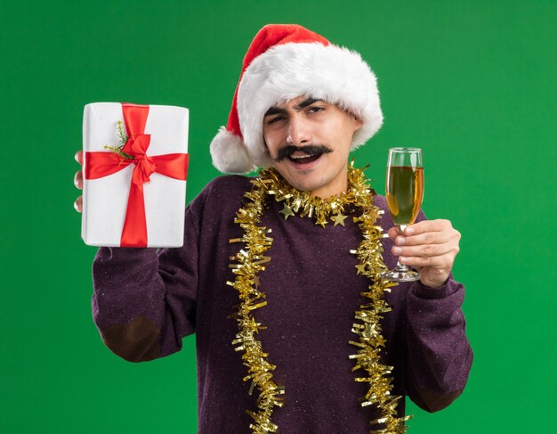 幸せな若い口ひげを生やした男は彼の首に見掛け倒しのクリスマスサンタの帽子をかぶってシャンパンとクリスマスプレゼントのガラスを保持している緑の背景の上に元気に立って笑顔のカメラを見て