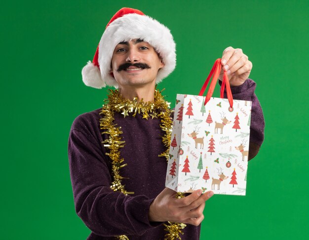 Happy  young mustachioed man wearing christmas santa hat with tinsel around his neck holding paper bag  with christmas gift looking at camera smiling confident standing over green background