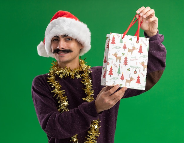 happy young mustachioed man wearing christmas santa hat with tinsel around his neck holding paper bag with christmas gift looking at camera smiling confident standing over green background