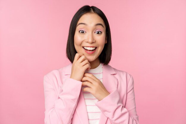 핑크색 스튜디오 배경에 포즈를 취한 카메라에 놀란 표정으로 양복을 입은 행복한 젊은 사무실 여성 한국 비즈니스 여성
