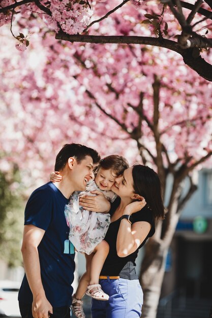 Happy young parents with a little daughter stand under blooming pink tree outside