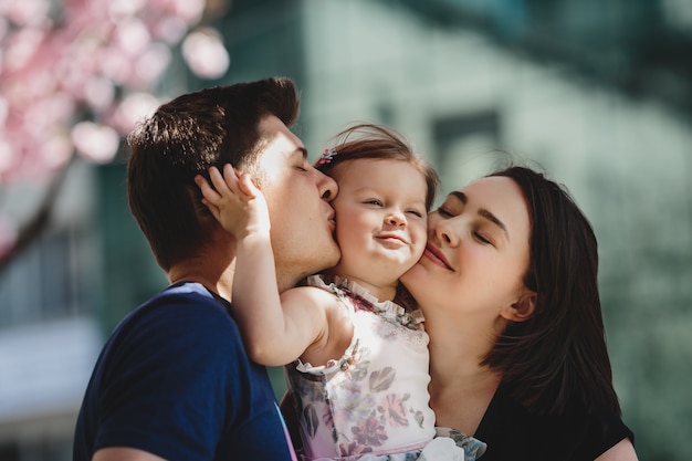 Happy young parents with a little daughter stand under blooming pink tree outside