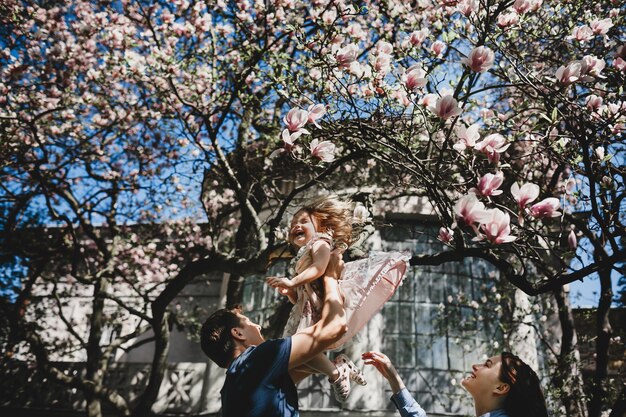 Happy young parents with a little daughter stand under blooming pink tree outside