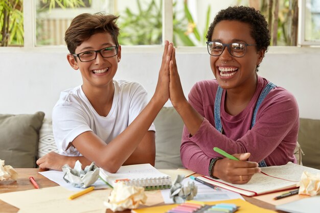 Happy young partners cooperate together in workspace, give high five to each other, have overjoyed expressions, work on research, write down records in notepad, ponder on idea, work as team.